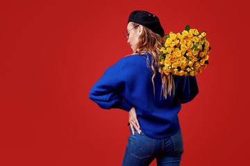 Cheerful young lady in blue clothes, black beret and round glasses being excited to get bouquet of yellow roses on women's day or birthday isolated over red background. French style girl 