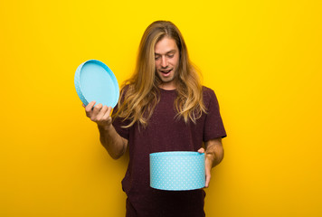 Wall Mural - Blond man with long hair over yellow wall holding gift box in hands