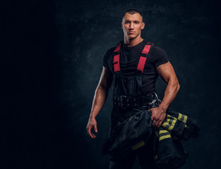 Wall Mural - A brave fireman holding a jacket and looking at a camera. Studio photo against a dark textured wall 