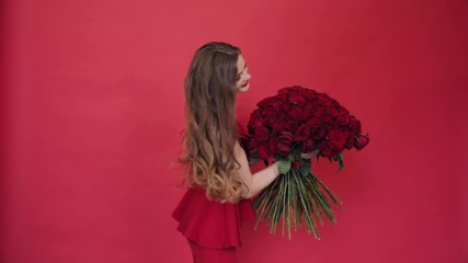 Wall Mural - Gorgeous woman in red dress and hairstyle spinning with big bouquet of red roses on isolated background. Pretty female enjoying gift and showing flowers in studio. Concept of present and beauty.