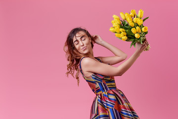 Cheerful young lady in stylish fashionable dress being excited to get bouquet of spring yellow flowers tulips on women's day isolated over pink background. 