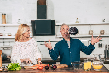 cheerful woman cutting carrot and looking at red cabbage in air near husband