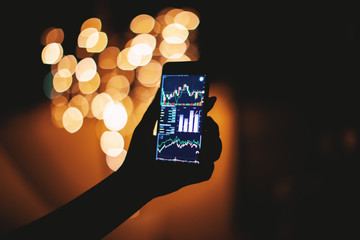 Silhouette of female hand holding smart phone in the dark with stock trading on display with light bokeh blurred background, Mobile business concept