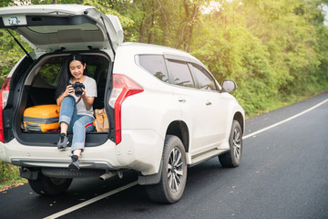 Woman traveler and camera sitting on hatchback car with natural background . Young traveler  woman taking a photo and sitting on hatchback car .