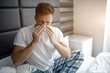 © estradaanton - Young man sit on bed early morning. He sneeze into white napkin. Sick and ill young man.