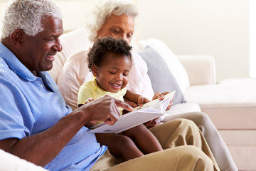 Wall Mural - Grandparents Sitting On Sofa At Home With Baby Granddaughter Reading Book Together