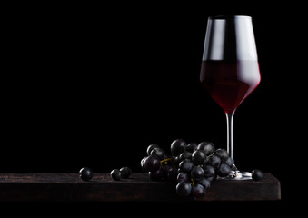 Elegant glass of red wine with dark grapes on wooden board on black background. Space for text