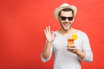 Portrait of attractive young man in hat and sunglasses standing and drinking orange juice over red background. Ok sign.