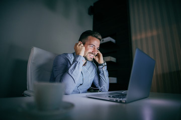 Smiling Caucasian employee preparing for conference call and putting earphones in ears while sitting in the office late at night.