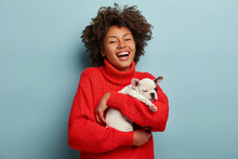 You Are My Friend! Pleased Laughing Young Woman With Gentle Look Embraces Sleepy Cute Puppy, Expresses True Love, Cares Of Favourite Pet, Dressed In Red Jumper, Isolated Over Blue Background