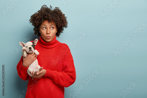 Dreamy beautiful woman with Afro hairdo, holds her best friend in hands, focused upwards, has so cute and lovable puppy, smiles while embraces pet, isolated over blue background with mock up space