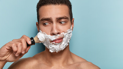 Wall Mural - Puzzled naked man has foam on face, keep gaze aside, shaves bristle, wants to have smooth skin, stands with bare shoulders against blue background, blank space on right side. Skin care concept