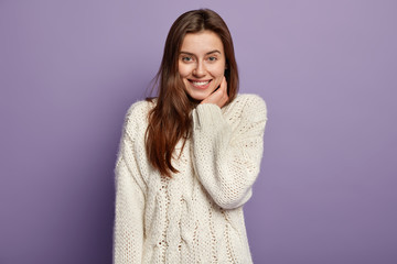 Wall Mural - Portrait of tender satisfied model bites lower lip, has toothy smile, keeps hand near cheek, expresses positiveness, hears pleasant news, dressed in white jumper, isolated over purple studio wall