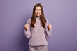 Gorgeous smiling young woman points down on floor, wears knitted sweater and corduroys trousers, advertises new shoes she bought in shop, isolated over purple backgroud. Advertisement concept