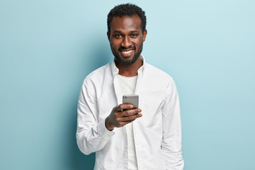 Ethnic man recieves email about discounts, uses application for publication, holds modern cell phone, dressed in white shirt, connected to wireless internet, waits for sms message, smiles positively