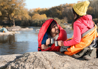 Sticker - Couple of campers in sleeping bags sitting on rock near pond. Space for text