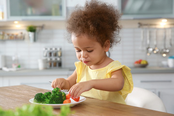 Cute African-American girl eating vegetables at table in kitchen