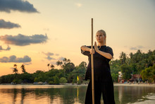 Young Beautiful Girl Woman Blond Doing Kung Fu With Bamboo Stick On The Seashore At Sunset