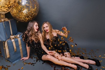 Wall Mural - Blonde girl sitting on the floor with friend and throwing out golden confetti on dark background. Stylish ladies in black dresses lying beside presents and balloons and joking.