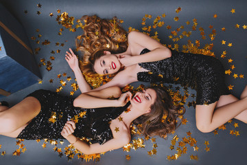 Overhead portrait of two joyful girls lying on golden confetti. Long-haired lady in black dress having fun with brunette sister at new year party.