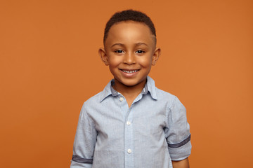 Wall Mural - Handsome adorable dark skinned schoolboy wearing neat blue shirt smiling broadly, rejoicing at good mark at school, posing against orange wall background with copy space for your advertising content