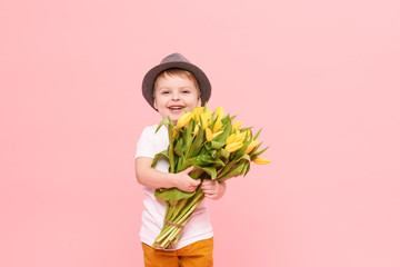 Adorable smiling child with spring flower bouquet looking at camera isolated on pink. Little toddler boy in hat holding yellow tulips as gift for mom. Copy space for text on right side