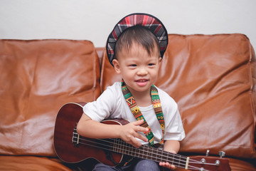 Cute happy smiling little Asian 2 - 3 years old toddler baby boy child wearing hat hold & play Hawaiian guitar or ukulele in living room at home, Music for kids and toddlers concept