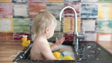 Wall Mural - Cute smiling baby taking bath in kitchen sink. Child playing with foam and soap bubbles in sunny kitchen with rubber ducks and toys. Little boy bathing, fun with water