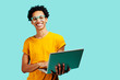 © Carlos David - Portrait of a smiling young man with glasses holding his laptop, isolated on blue background
