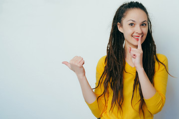 Cheerful dreadlocks-haired woman laughing out loud pointing finger at funny advertisement copy space aside, says shh on white grey studio background