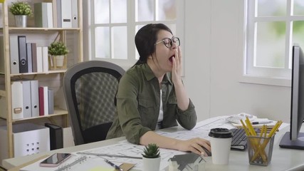 Wall Mural - tired girl interior designer ready to work in morning sitting at desktop with working tools. young lady architect yawning hand covering mouth using computer with blueprint and coffee on table.