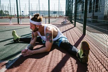 Wall Mural - Athletic guy with white headband, dressed in the white t-shirt, black leggings and blue shorts doing stretching on the playground outside on a sunny day