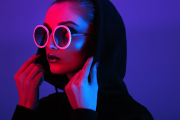 Portrait of fashion young girl in a black sweater with a hood and round sunglasses in red and blue neon light in the studio