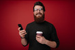 © Vulp - Handsome bearded man holding coffee and phone in hands. Hipster bearded man looking at camera while holding mobile and coffee