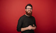 © Vulp - Image of happy young bearded man standing over red wall background isolated. Looking camera using smartphone