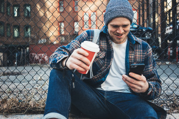 Wall Mural - Handsome man sitting on the city street resting with cup of hot coffee and using mobile phone