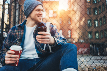 Wall Mural - Smiling man sitting on the city street resting with cup of hot coffee and using mobile phone