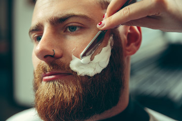 Wall Mural - Client during beard shaving in barbershop. Female barber at salon. Gender equality. Woman in the male profession. Hands close up