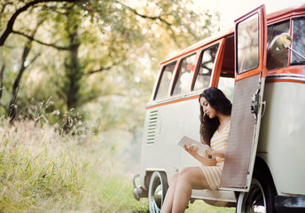 Wall Mural - A young girl with a book by a car on a roadtrip through countryside, reading.