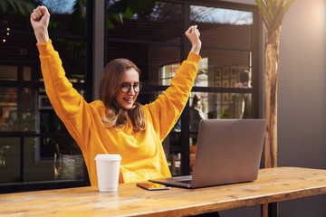 Girl rejoices in winning having raised her hands up sitting in front of laptop.Woman watches sports match on computer.