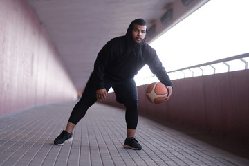 Stylish African-American guy wearing a black hoodie playing basketball on a bridge sidewalk 