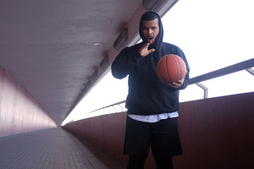 Wall Mural - Stylish African-American guy wearing a black hoodie holding a basketball and yawning while standing on a bridge sidewalk