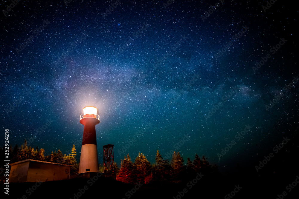 A lighthouse at night with a star filled sky above. The light in the top of the lighthouse is illuminated. The Milky Way is visible. - obrazy, fototapety, plakaty 