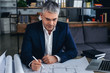 © Pixel-Shot - Mature architect working with drawings in office