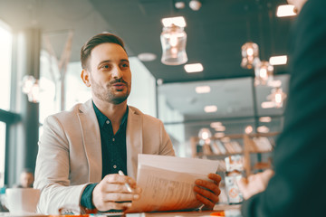 Positive handsome Caucasian unshaven businessman holding paperwork and talking to his colleague while sitting in cafeteria. Work so hard that one day your signature will be called an autograph.