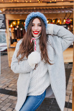 Laughing Female Model With Dark-brown Hair Eating Christmas Sweets In Cold Day. Portrait Of Merry Brunette European Girl In Gray Coat And White Mittens Posing With Lollipop In Winter Morning.