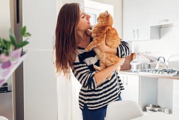 Wall Mural - Young woman playing with cat in kitchen at home. Girl holding and hugging ginger cat