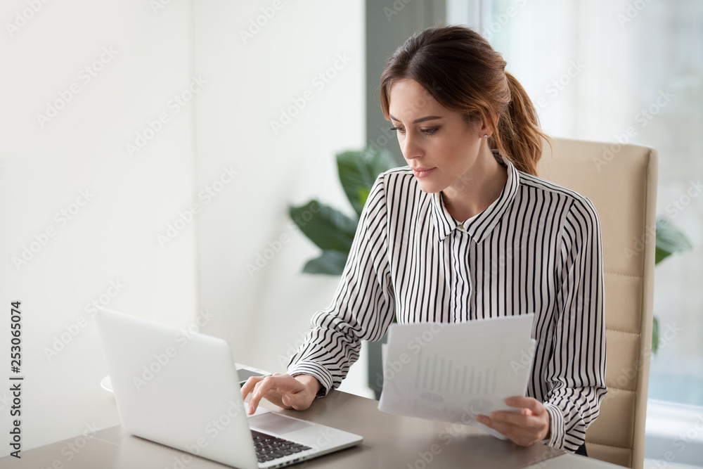 Serious focused businesswoman typing on laptop holding papers preparing report - obrazy, fototapety, plakaty 