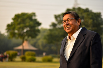 Wall Mural - Happy looking senior Indian man wearing a suit standing and smiling, laughing in a park in Delhi, India