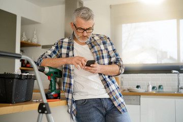 Wall Mural - Mature man checking phone whilst fixing shelf in modern kitchen